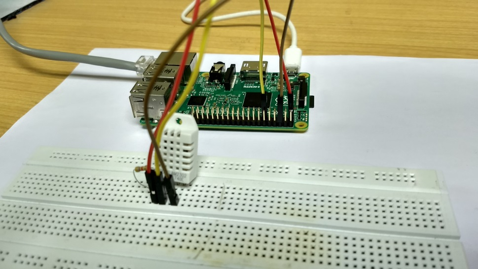 Photograph showing a DHT22 temperature and humidity sensor (white plastic housing with ventilation slots) connected to a Raspberry Pi GPIO header via jumper wires on a breadboard. The setup demonstrates proper wiring for the popular DHT22 sensor: VCC to 3.3V power, GND to ground, and DATA pin to a GPIO pin with a pull-up resistor for reliable 1-wire communication.
